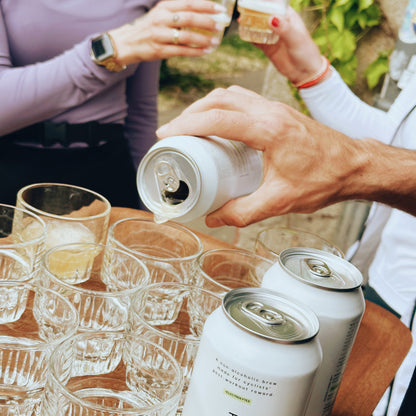 Person pouring beer from a can into glasses with another person clinking glasses in the background.
